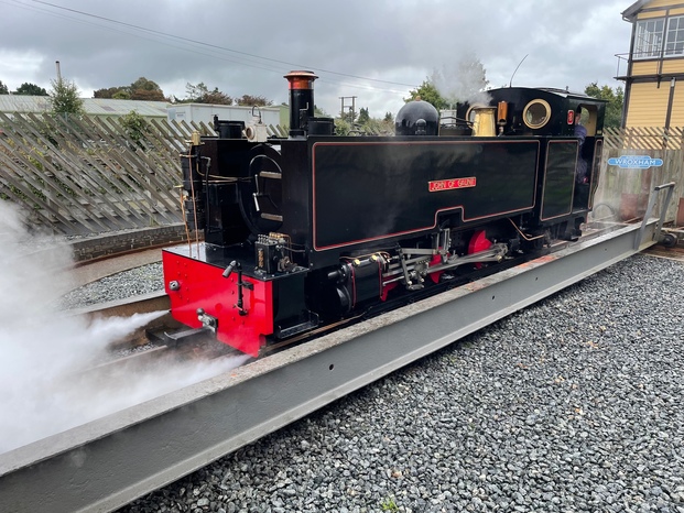 A black steam engine sitting on a turntable, blowing steam out of several pipes and holes. It’s facing towards the camera – it’s just been turned around from the other direction.